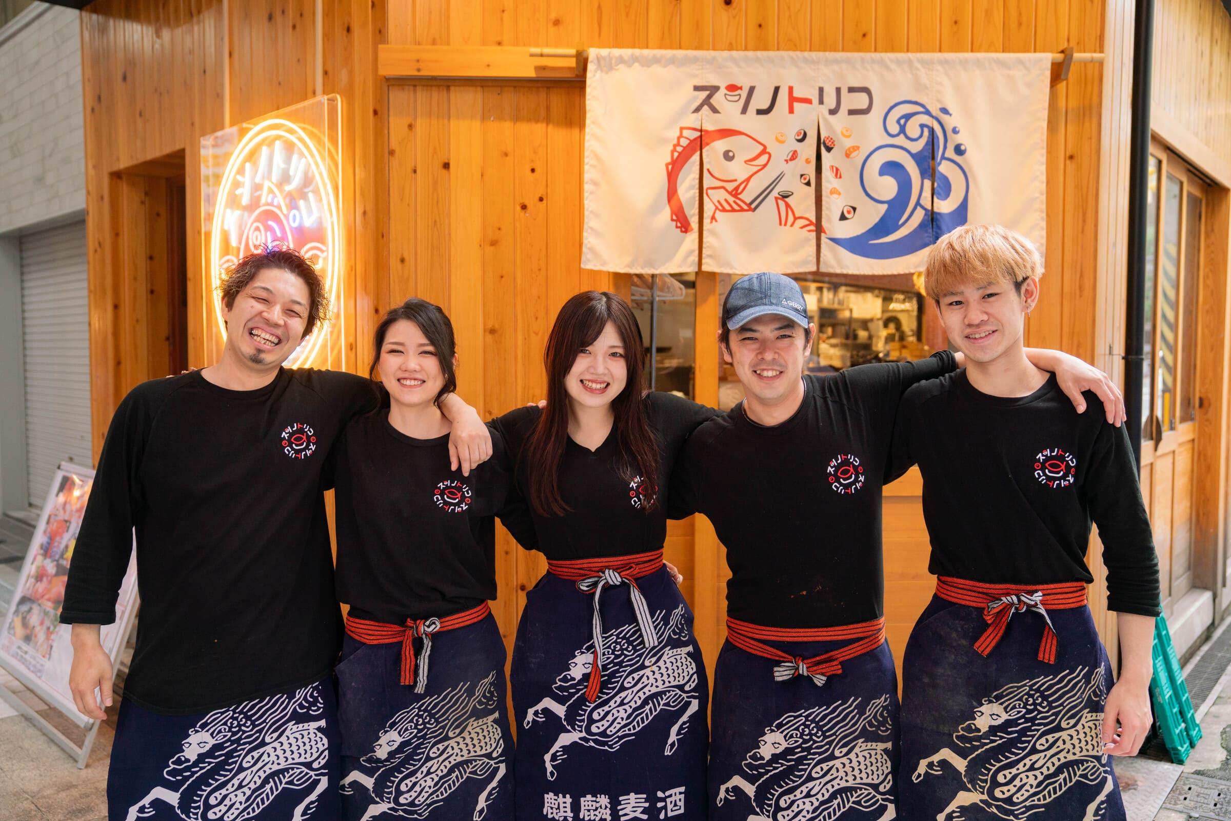 スシノトリコのスタッフが、店の前で笑顔で肩を組んでいる様子